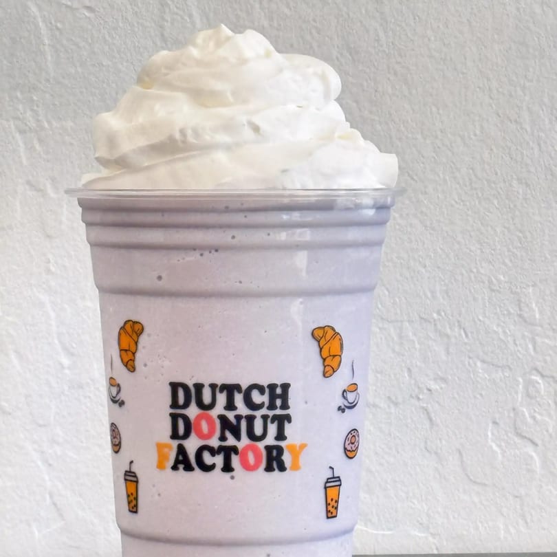 A Dutch Donut Factory branded cup filled with a light purple drink and topped with whipped cream.