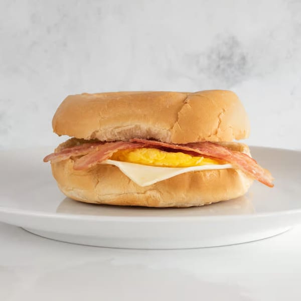 A breakfast sandwich with bacon, egg, and cheese on a bun, served on a white plate.