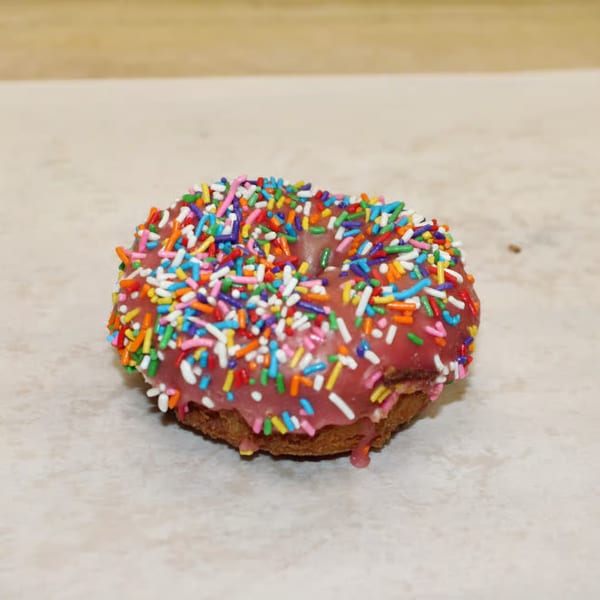 A single pink-frosted donut with colorful sprinkles sits on a light-colored surface.