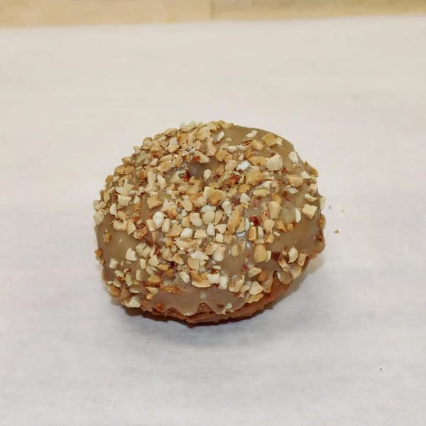 A Dutch Donut Factory donut with light brown glaze and chopped nuts on a white surface.